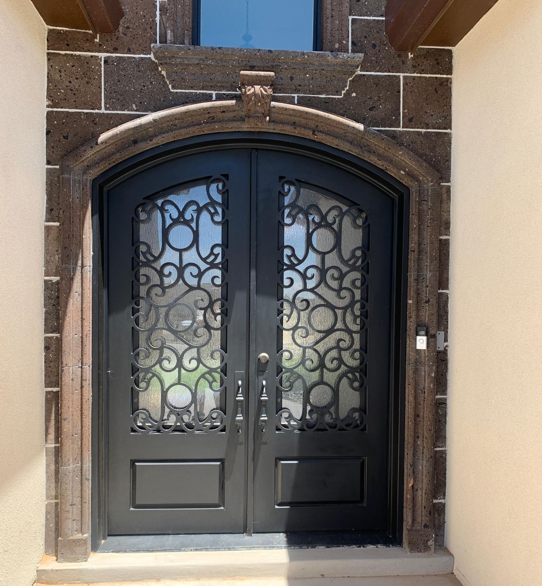 Black wrought iron arched double entry door with scrollwork and rain glass, installed in a stone surround